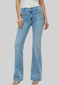 Jeans acampanados de color azul claro, hechos de denim, con bolsillos frontales con cremallera y herrajes en color dorado, y un diseño de cintura alta.