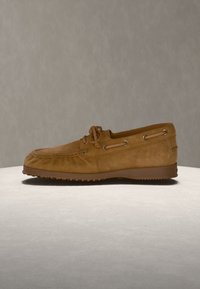 Mocassin marron en daim style mocassin avec détail de lacet et semelle en caoutchouc sur une surface claire et fond neutre.