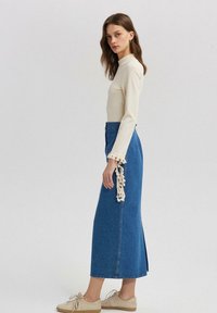 Lichtbeige blouse met lange mouwen en decoratieve kantafwerking, gecombineerd met een blauwe denim maxirok met zijsplitsen en subtiele versieringen.