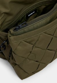 Borsa verde oliva trapuntata con tasca con zip, superficie tessurizzata e hardware della cerniera nera. L'interno ha una rifinitura liscia e l'etichetta ben visibile.