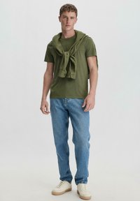 Ein olivgrünes T-Shirt mit Rundhalsausschnitt, unterlegt mit einem Pullover, der um die Schultern gebunden ist, kombiniert mit hellblauen Jeans und cremefarbenen Sneakern.