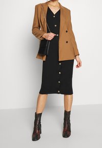 Braunes, doppelt knöpfbares Blazer über einem schwarzen, gerippten Kleid mit goldenen Knöpfen. Schwarze Handtasche mit Kettenriemen und gemusterten Stiefeletten.