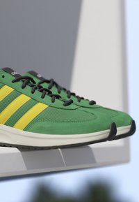 Green Adidas sneakers with yellow stripes and black laces resting on a white surface against a minimal gray and blue background.