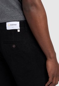Schwarze Cordhosen mit strukturiertem Finish, ausgestattet mit einem braunen Knopf, Seitentaschen und einem weißen Etikett mit dem Markennamen "Farah."