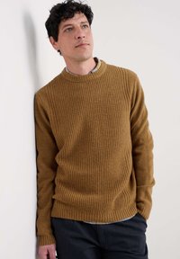 Brauner Strickpullover mit strukturierten Oberflächen und gerippten Bündchen, kombiniert mit einem karrierten Hemd, vor einem schlichten Wandhintergrund.