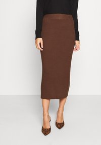 Femme portant une jupe midi marron côtelée ajustée et des escarpins à bout pointu imprimé léopard, debout devant un fond uni.