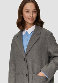 Grijze wolmix jas met een notch-kraag, twee voorzakken en zwarte knopen, gelaagd over een lichtblauw overhemd met kraag.