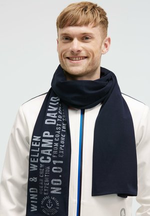 Smiling man with short hair wearing a navy blue scarf with white text over a white jacket with blue zipper accents.