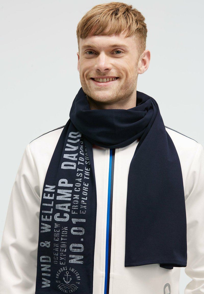 Smiling man with short hair wearing a navy blue scarf with white text over a white jacket with blue zipper accents.
