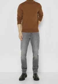 Brun rullekravetrøje, grå jeans og sorte ankelstøvler. Trøjen har ribbede manchetter og kant, jeansene har lige ben.