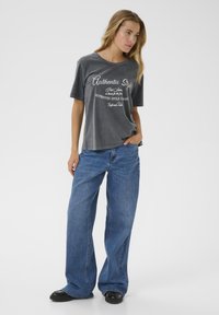 T-shirt gris surdimensionné avec un texte graphique blanc à l'avant, associé à un jean bleu à jambe large et des chaussures noires. Tissu lisse, style décontracté.