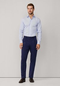 Uomo in piedi con camicia azzurro chiaro, pantaloni a pieghe blu navy e scarpe eleganti marroni scure su uno sfondo grigio semplice.