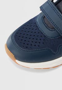 Κοντινό πλάνο στα δάχτυλα ενός ναυτικού μπλε αθλητικού παπουτσιού με διάτρητο πλέγμα, δερμάτινο λουράκι, λευκή μεσαία σόλα και ελαφρώς καφέ λαστιχένια εξωτερική σόλα.