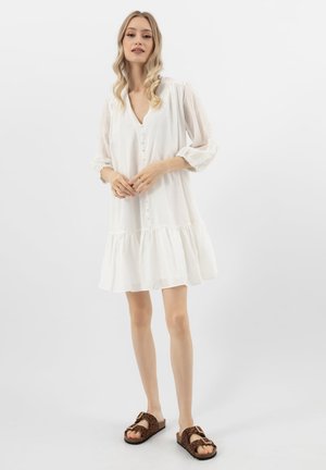 Frau steht in einem weißen, geknöpften Kleid mit Puffärmeln und Sandalen mit Leopardenmuster auf Schnallen vor weißem Hintergrund.