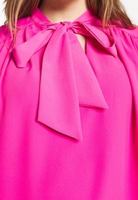 Fuksia-paita, jossa on rusetti yksityiskohtana kaula-aukossa, valmistettu tekstuurisesta kankaasta, jossa on sileä viimeistely ja lyhyet hihat.