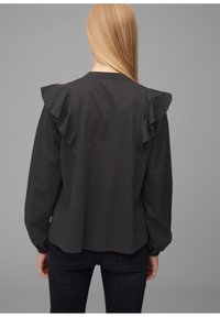 Black long-sleeve blouse with ruffled shoulder accents, relaxed fit, and smooth texture. Worn with dark jeans.