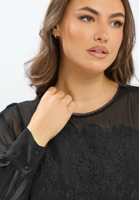 Femme aux cheveux bruns ondulés portant un blouse en dentelle noire avec des manches transparentes, tenant le col près du cou, regardant sur le côté avec une expression neutre.