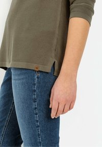 Osoba w niebieskich dżinsach i oliwkowozielonej, strukturalnej bluzce z krótkim rękawem i bocznym rozcięciem, stojąca na jednolitym tle.