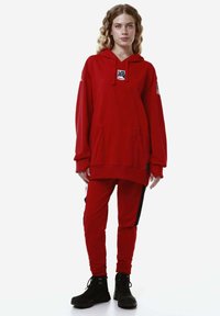 Felpa rossa con tasca frontale e logo, abbinata a pantaloni jogger rossi con pannelli laterali neri e stivali neri con lacci.