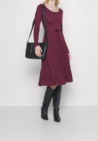 Robe à manches longues de couleur bordeaux avec une jupe plissée. Associée à un sac bandoulière noir et des bottes noires montant jusqu'aux genoux. Design simple, texture douce.