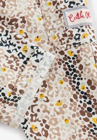 Gros plan d’un tissu beige avec des taches de léopard marron et noir ainsi que de petites fleurs jaunes, montrant une poche cousue et une étiquette avec un texte rouge.