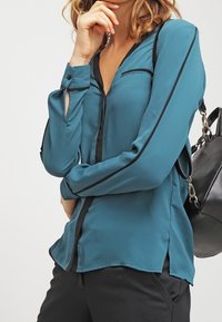 Turkos långärmad blus med svarta detaljer, knappade ärmslut och V-ringning. Har sidoflikar och en mjuk textur. Stylerad med en svart väska.