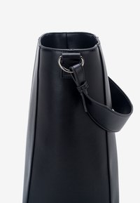 Sac à main en cuir noir avec une large bandoulière attachée par un anneau métallique argenté, présenté de côté sur un fond uni clair.