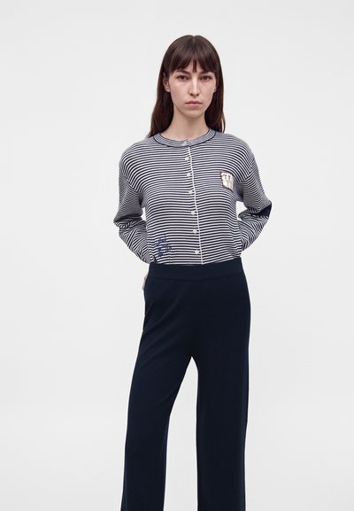 Femme aux longs cheveux bruns porte un haut boutonné rayé noir et blanc et un pantalon noir, debout les mains derrière le dos.