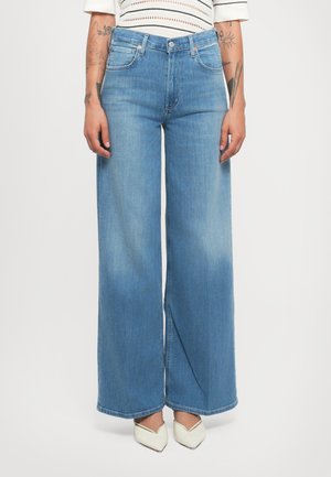 Personne portant un jean large bleu clair, des chaussures blanches à bout pointu et un top court rayé blanc, avec des tatouages visibles sur les bras.