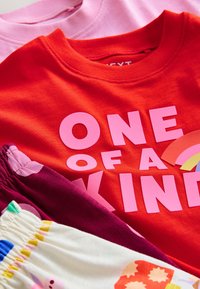 Červené tričko s růžovým textem "ONE OF A KIND" a duhovou grafikou, vedle růžových, vínových a vzorovaných kusů látky.