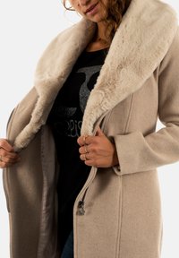 Beige uld-blandede frakke med en fuzzy cremet kunstpelskrave, lynlås lukning og sidelommer. Båret over en sort grafisk t-shirt.
