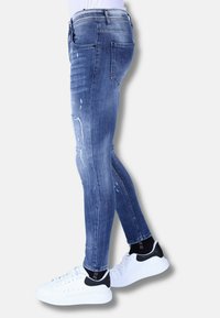 Blauwe skinny jeans met een distressed ontwerp met scheuren, een standaard tailleband, gecombineerd met witte sneakers en zwarte sokken.