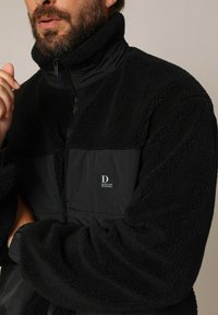 Zwarte fleece jas met hoge kraag, voorzien van een gladde zwarte nylonsectie, logo patch en textiel voor warmte en comfort.