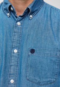 Muž oblečený v modré džínové košili s knoflíky, která má na hrudi kapsu a tmavě vyšívané logo v levém horním rohu kapsy.