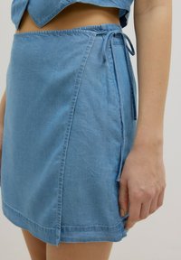 Hellblauer Chambray-Wickelrock mit Kordeldetail, strukturiertem Stoff, sauberen Nähten und geradem Saum, der einen seitlichen Schlitz aufweist.