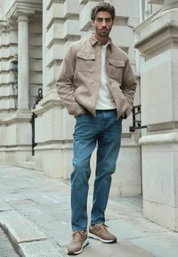 Jachetă din suede bej, cămașă tricotată albă, blugi albaștri și adidași maro. Modelul stă relaxat pe un drum de piatră, în fața unui perete texturat.