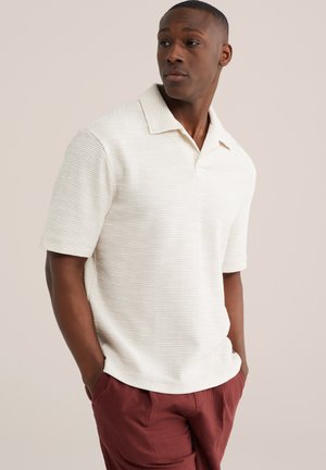 Mann trägt ein strukturiertes weißes Polo-Shirt mit kurzen Ärmeln und rostfarbene Hose, steht mit Händen in den Taschen und schaut zur Seite.