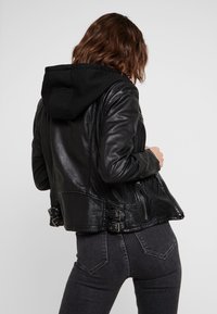 Veste en cuir noir avec une capuche détachable noire, design ajusté, poches zippées et accents en boucle à la taille. Fini texturé.
