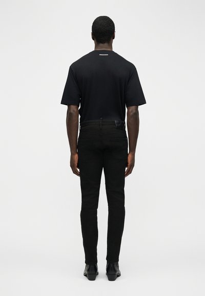 Άνδρας που στέκεται γυρισμένος από την πλάτη, φορώντας μαύρο κοντομάνικο πουκάμισο και μαύρο στενό παντελόνι με μαύρα παπούτσια, σε λευκό φόντο.