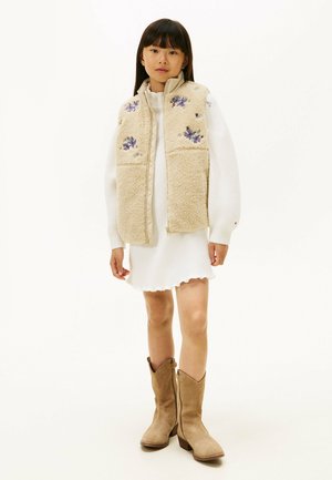 Jong meisje staand, gekleed in een witte gebreide jurk, een beige fleecevest met geborduurde bloemen en lichtbruine suède laarzen tot halverwege de kuit, tegen een witte achtergrond.