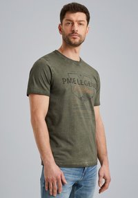 T-shirt en coton vert olive avec un col rond, des manches courtes et un design graphique à l'avant comportant du texte et des embellissements.
