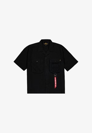 Kratke rokave črna majica iz tekstila, ki ima dva sprednja žepa, zasnovo na gumb in rdečo nalepko z besedilom ter kovinsko sponko.