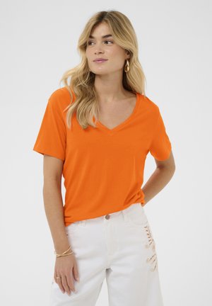 Žena s dlhými blond vlasmi, ktorá nosí zlaté kruhové náušnice, oranžové tričko s výstrihom do V a krátkymi rukávmi a biele nohavice so zaväzovacím detailom.