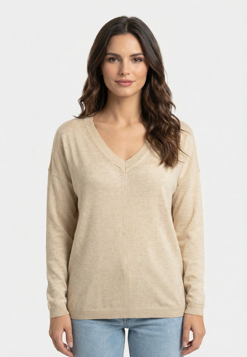 Femme aux longs cheveux bruns ondulés, portant un pull beige à encolure en V et un jean bleu clair, debout devant un fond blanc uni.
