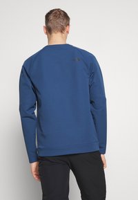 Uomo di spalle che indossa una felpa blu a maniche lunghe e pantaloni neri su uno sfondo chiaro e uniforme.