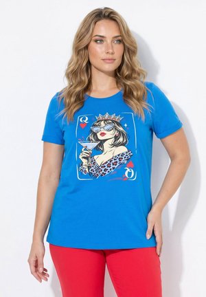 Donna con capelli biondi mossi che indossa una maglietta blu con un'illustrazione della Regina di Cuori e pantaloni rossi, in piedi con una mano sui fianchi.