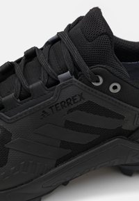 Czarne sportowe buty z siateczkową cholewką i syntetycznymi wstawkami. Posiadają teksturowane detale, system sznurowania oraz wyraźne logo marki.