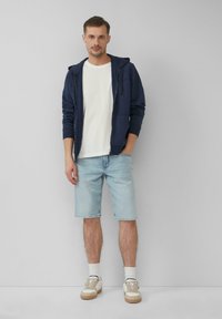 Un sweat à capuche zippé marine, un t-shirt blanc, un short en denim bleu clair et des baskets blanches avec des accents beiges, se tenant devant un fond neutre.
