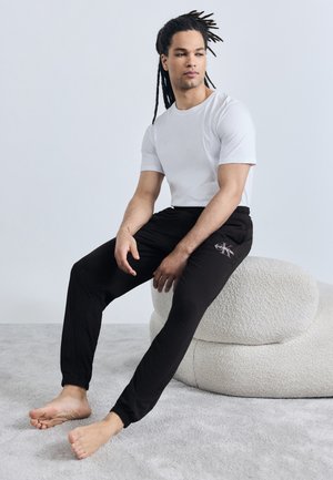 Άνδρας με μακριά dreadlocks που φοράει λευκό μπλουζάκι και μαύρη φόρμα Calvin Klein, καθισμένος ξυπόλυτος σε ένα λευκό μαξιλάρι με υφή.