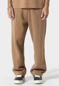 Ruske sweatpants s opuštenim krojem, vidljivim šavovima i glatkom teksturom, upareni s crnim tenisicama i bijelim vezicama.